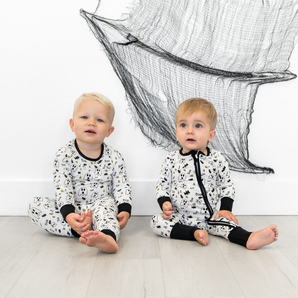 Two children in black and white patterned outfits sitting on a light wood floor with a decorative net in the background.