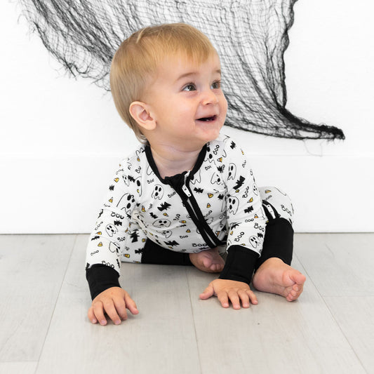 Baby wearing a black and white patterned onesie sitting on a light-colored floor with a white wall and black net in the background.